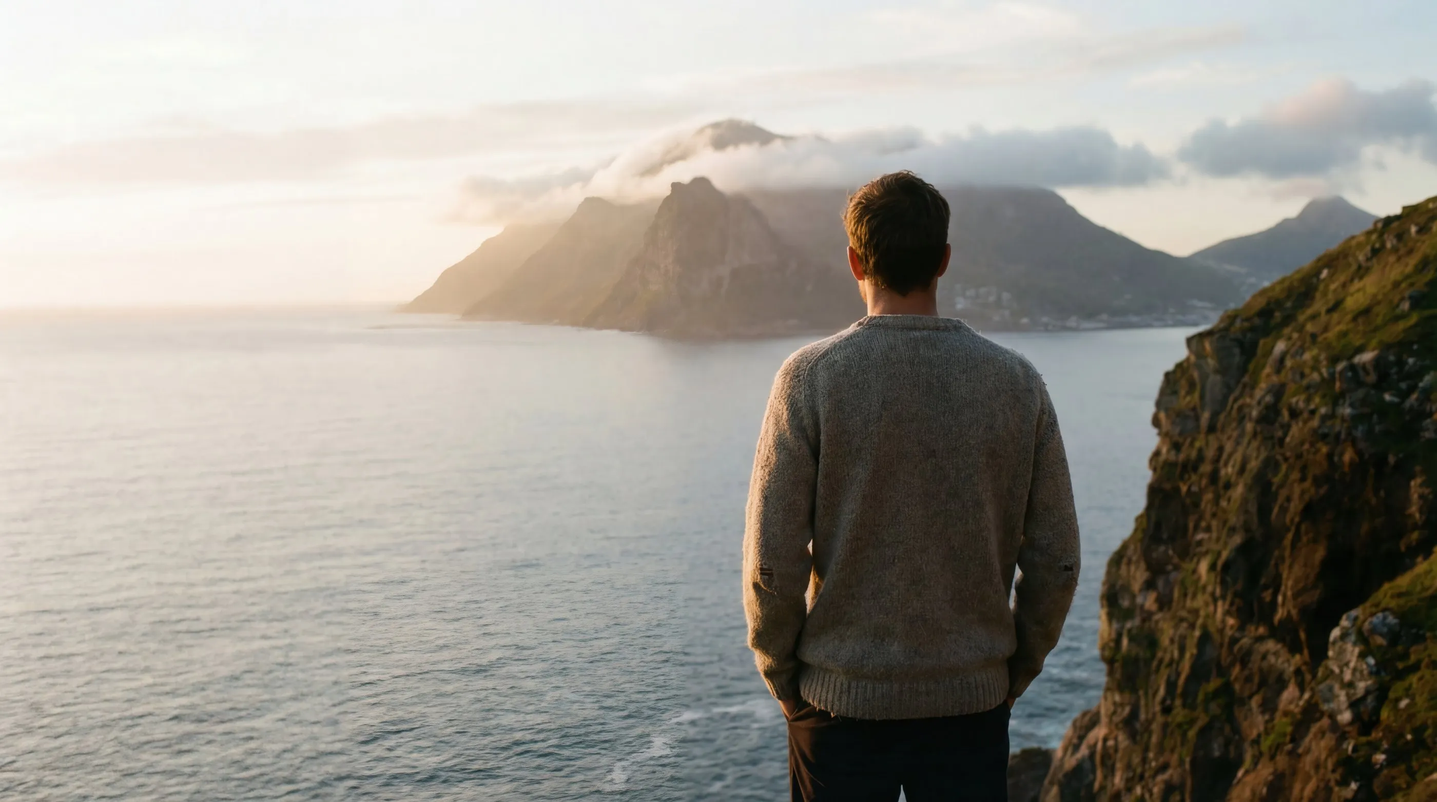 A man seen from behind, looking out over the sea toward misty mountains at sunrise